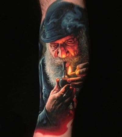 Tatuagem realista de um homem fumando um cachimbo, com um palito de fósforo acesso na mão.